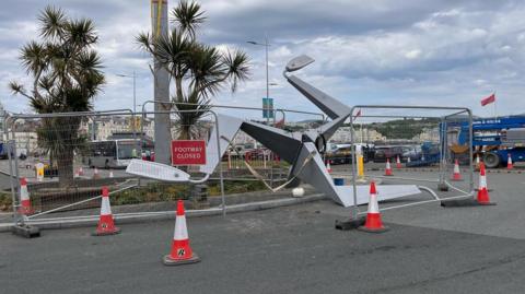 A large steel structure of a triskelion on part of a roundabout ahead of it being mounted. The structure is surrounded by fencing and traffic cones. 