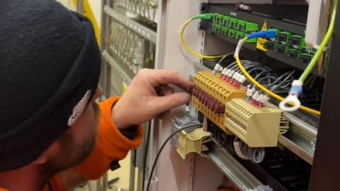 A worker wearing a black beanie and an orange high-vis uniform is working on the wires in a signalling box. 