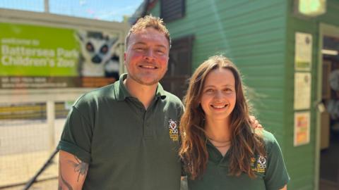 A man with blonde hair and a woman with long brown hair, both wearing green polo shirts, smile into the camera in front of Battersea Park Children's Zoo