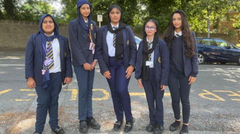 A group of five schoolgirls in dark blue uniforms standing on a roadside next to a yellow 'keep clear' sign