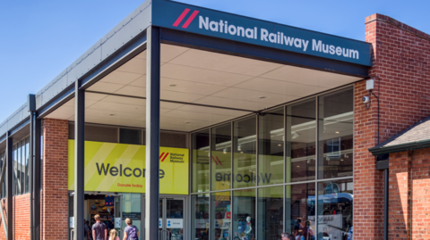 The exterior of the National Railway Museum on a sunny day. The museum building is red brick with blue and red signs and a bright yellow welcome sig above the entrance.  Two adults and a child are walking into the museum. 