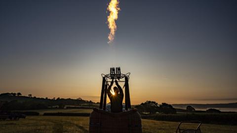 A person standing in a hot air balloon basket fires up the burners against an early morning sky of dark blue and orange