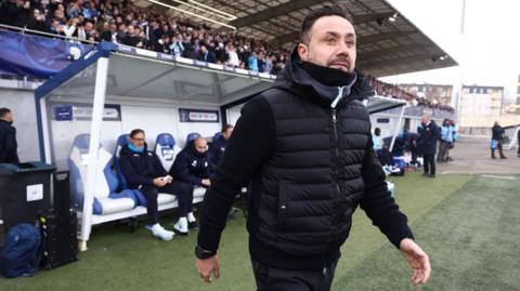 De Zerbi, a man wearing a black puffer jacket with a long-sleeved black top underneath, walking on to a football pitch