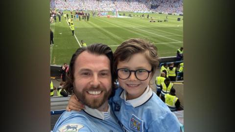 A picture of Matt Bradley on the left smiling at the camera wearing a sky blue Coventry City top and his son on the right smiling at the camera wearing glasses and a sky blue Coventry City top. Behind is the CBS Arena with the green football pitch, fans far in the opposite stands and stewards dotted around.