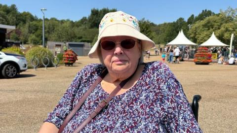 There is a woman in a white bucket hat, which has flowers on. She is wearing oval thin-framed sunglasses and a blouse which has dark blue, dark purple and white splodges on. She has a brown strap for her handbag. She is also sat in a wheelchair.