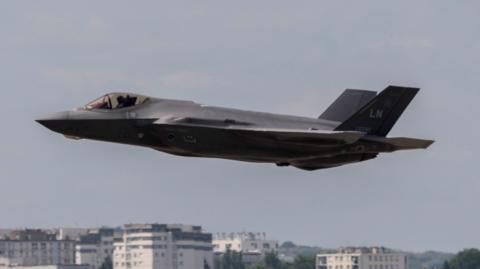A fighter jet in the air against the sky, with the tops of some buildings visible in the background