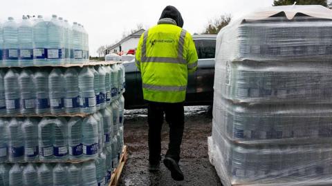 A person in a yellow high vis coat with "South East Water" written on the back. They are walking between two crates of bottled water.