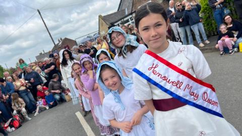 A group of children dressed in traditional parade clothing walk along a village street. The child at the front wears a white outfit with a sash reading "Hemswell May Queen" and is followed by others in white clothing and patterned headscarves. A large crowd of spectators lines the roadside behind them.