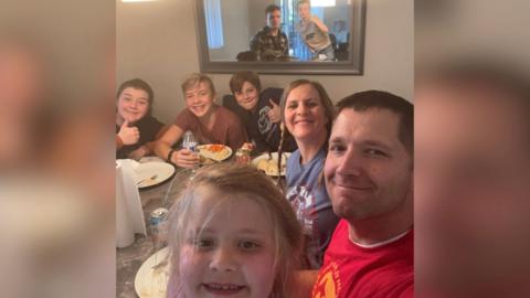 A man, woman, and their six children sitting around a dining table having just eaten a Thanksgiving meal.