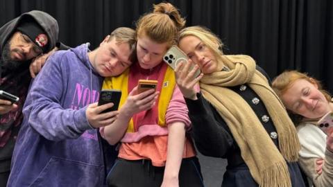 The cast of Generation Fomo stand together looking at phones.