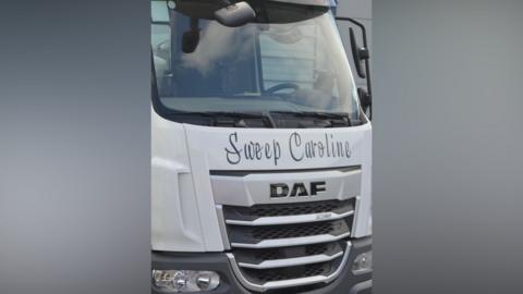 A close-up photograph of the front of what appears to be a white light goods vehicle/lorry. "Sweep Caroline" can be read on the front in black text.