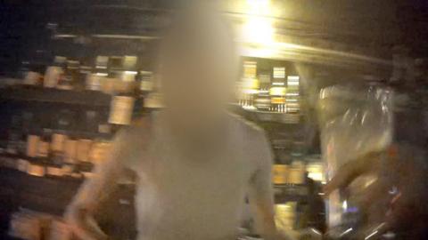 Blurred image of a man at a bar at night taken with a secret camera. In the foreground a hand is holding a glass, in the background there are bottles on shelves.