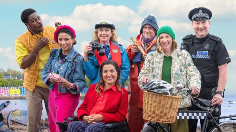 The cast of Balamory, all dressed in colourful clothes, smile at the camera 