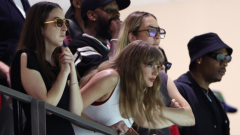 Taylor Swift, wearing a white vest top, leans over some railings at the Super Dome in New Orleans