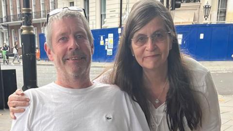 Andrew and Joanna Chadwick pose together for a photo outside. They are on a street in front of a large blue hoarding in front of a stone building. Andrew has short white hair and a beard with his glasses on his head and a white t-shirt. His wife has her right arm round he shoulders and has long dark hair and glasses