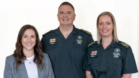 Heather Langlois, John Atkins and Aimee Lihou. Stood facing the camera with a white background. John and Aimee have St John dark green uniforms on and Heather is wearing a grey suit jacket and white top. 