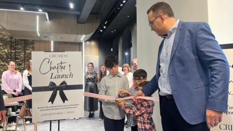 Three small boys cut a ribbon in a shop with a man in a suit - their father - assisting. A sign says Creaseys Chatper 1 Launch - 20 September 2025.