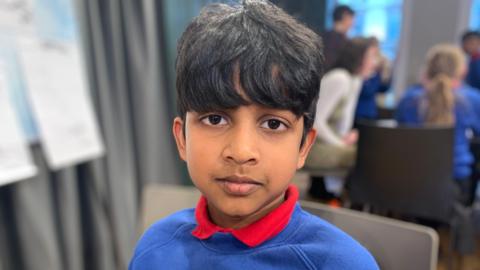 A boy with dark hair is looking at the camera. He is wearing a blue sweatshirt with a red t-shirt underneath. There are children sitting at tables in the background.