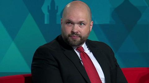 Peter Lamb, a bald man with a beard and moustache, sits on a red sofa in the BBC South East studio. He is wearing a black suit, red tie and white shirt.