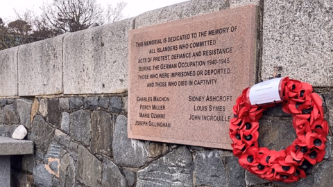 A plaque dedicated to the Guernsey Eight, with a wreath of poppies next to it.
