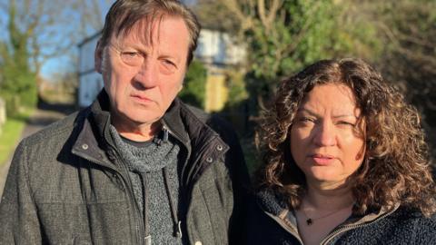 David and Karen Yates, pictured at Earls Farm in Bolton. David has short dark hair, and is wearing a dark grey jacket. He stands to the left of Karen, who has dark shoulder-length hair. Both look frustrated.