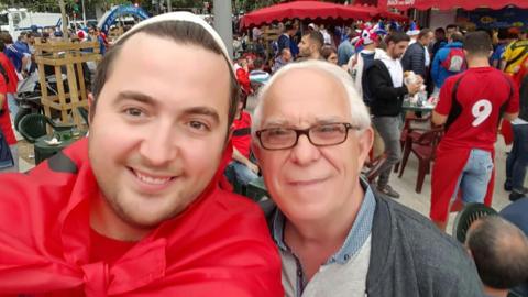 Hasan and his dad Emin pose for a photo. They are standing in a football fan zone.