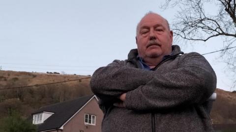 A bald man with a moustache wearing a grey fleece, looking angry with his arms crossed across his body. Behind him are houses and hills.