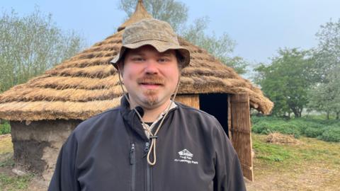 Arthur Randall looking at the camera. He is wearing a sunhat and a Flag Fen Archaeology Park-branded jumper. Behind him is the replica roundhouse, with layers of thatched roof over a circular wall. There is an open wooden door at the front. 