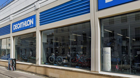 The side exterior of Decathlon, a sporting goods shop. There is blue and white branding above large windows, through which you can see lots of bikes arranged for sale.