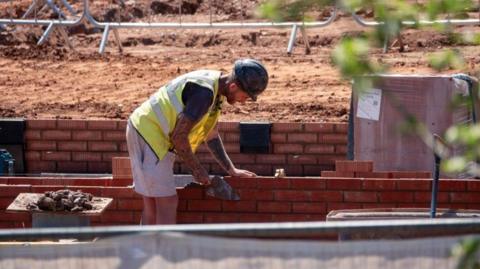 A builder wearing a hard hat, grey shorts and a hi-vis waistcoat over a dark coloured T-shirt is erecting a red brick wall on a building site.