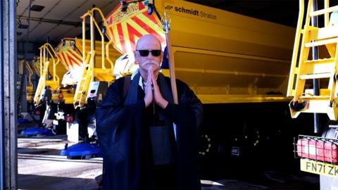 Zen Buddhist monk the Reverend Gando Seiko Garrod wearing a dark robe and a wooden staff with a metal emblem on top stands with his hands clasped in prayer in front of the bright yellow gritter lorries