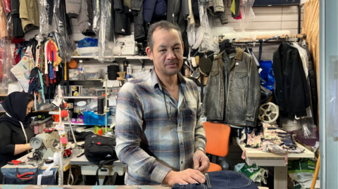 A man with brown hair and brown stubble on his face. He is wearing a blue, white and brown flannel and standing at a market stall which has clothing hung up on the wall behind him, and sewing equipment scattered around.  A woman with a head scarf on is sitting at a desk with a tape measure around her neck