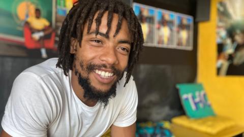 Donovan Charles Lansiquot sits in a community centre, smiling at the camera. There is bright artwork behind.