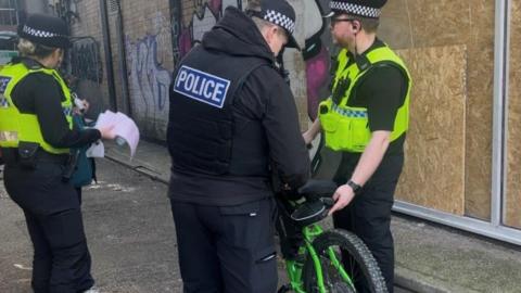 Three police officers standing in a street with two of the officers holding a green bike. The other officer is talking to a person stood in the street. There is graffiti on the walls in the background.