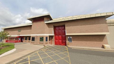 Outside of a prison with large brick walls and a red door for vans