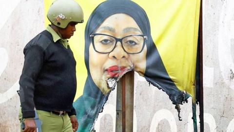 A Tanzanian riot police officer walks past a vandalised campaign poster of President Samia Suluhu Hassan showing her dressed in a black headscraf against a yellow background - 30 October 2025.