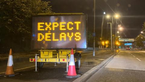 A black sign saying "expect delays" is lit up. It is on the side of the road and there are two traffic cones in front of it.
