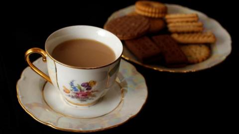 A cup of tea, on a saucer, and a plate of biscuits. The tea cup and plates are white, with a floral pattern, and in edged in gold. Chocolate and plain biscuits are on the plate.