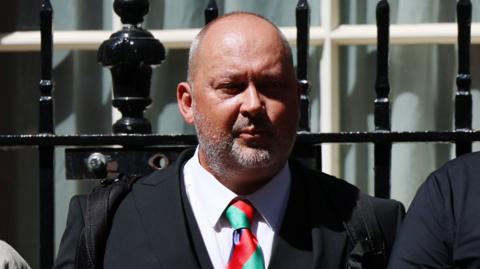 Olly Stephens' father Stuart stands outside 10 Downing Street. He is wearing a dark suit and a red and green tie.