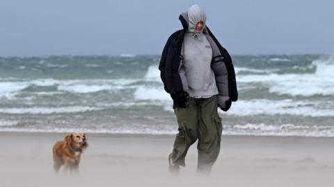 Man and his dog walking along a beach where sand is visibily lifted up with large waves in the background