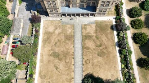 A parched lawn with historic patterns against a stately home