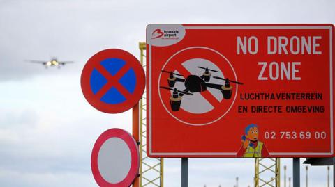 Photo shows a sign that reads 'No Drone Zone' near the Brussels-National Airport. A Brussels Airlines passenger jet can be seen coming in to land in the background.