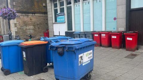 A view of communal bins outside of a closed shop. The windows have white paper in them, and a for sale sign to the left of the building. There are large communal bins on the left, and a line of bins against the shop.