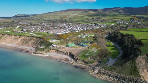 An aerial view of Glen Wyllin coast line and the village of Kirk Michael among green fields, ahead of a vast view of fields and rolling hills, on a sunny day.