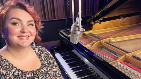 A woman with short, died red hair and a black floral dress, sat at a piano with a microphone in front of it. She is looking at the camera and smiling.