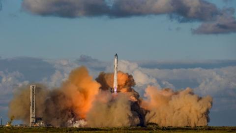 A rocket launches at the center of the image with plumes of smoke surrounding it. The sky in the background is blue with white and grey clouds.