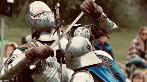 Two people in silver armour and chainmail are battling eachother and have their swords crossed. The one with their back to the camera is also wearing a blue silky cape. They have full visors and are wearing brown leather gauntlets.
Behind them, out of focus, are spectators.