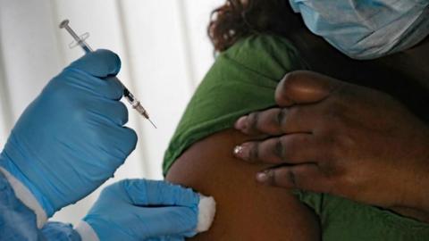 A person is shown receiving a flu vaccine in their upper right arm. A person can be seen holding a needle in a blue gloved hand and dabbing a spot on the patient's arm with a cotton bud. 