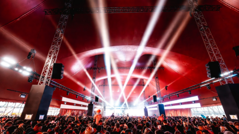 A packed crowd gathers inside a large red festival tent as bright white stage lights beam over the audience during a live music performance.