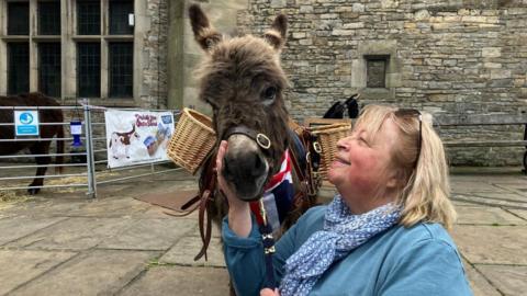 A woman with a short blonde bob, wearing a blue jumper and blue patterned scarf, holds the lead rope of a miniature donkey. The donkey is wearing a union jack neckerchief and a saddle with two baskets on either side. The woman is smiling and petting the donkey's nose. 